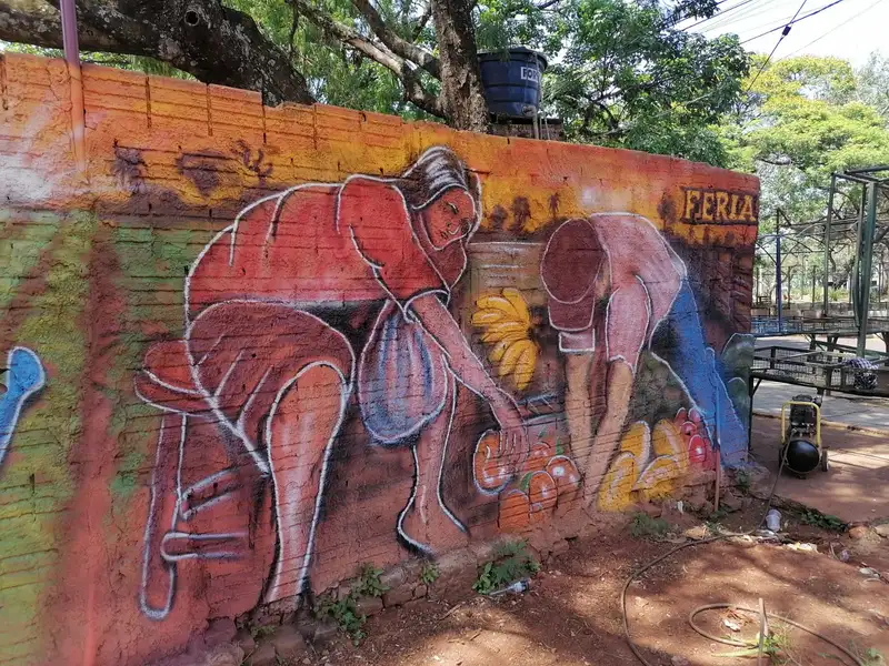 Un mural colorido de gran escala siendo pintado en el lateral de un edificio gris, transformando el entorno.